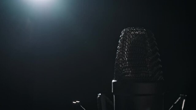 The studio condenser microphone in backlight rotates on a black background. Close-up of chrome grid of mic surface spinning around. Concept recording studio, voice, podcast, karaoke, audiobook. Copy