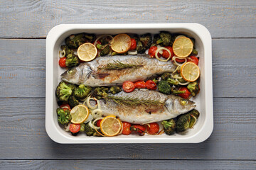 Delicious fish with vegetables and lemon in baking dish on grey wooden table, top view