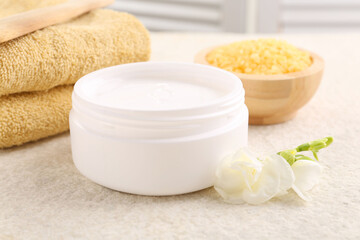 Fototapeta na wymiar Body care. Moisturizing cream in open jar and flower on light textured table, closeup