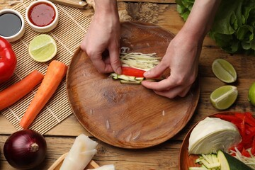 Making delicious spring rolls. Woman wrapping fresh vegetables into rice paper at wooden table, flat lay - obrazy, fototapety, plakaty