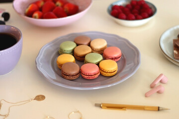 Obraz na płótnie Canvas Plate of pastel macarons, cookies and chocolate, cup of tea of coffee, glass of bubble water, various berries, books and accessories on the table. Selective focus, pastel colors.