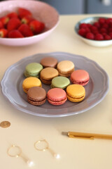Plate of pastel macarons, cookies and chocolate, cup of tea of coffee, glass of bubble water, various berries, books and accessories on the table. Selective focus, pastel colors.
