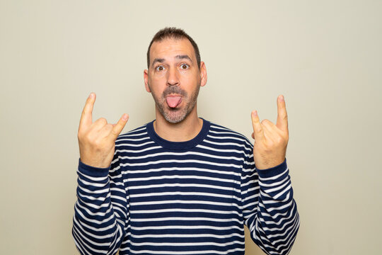 Portrait of a bearded man in his 40s wearing a striped sweater showing a rock and roll gesture with a heavy metal sign, enjoying his favorite music at the party. Isolated on beige background