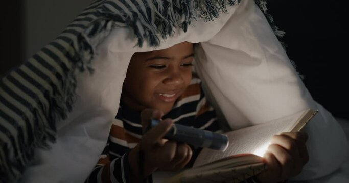 Adorable child reading book holding flashlight lying in bed under blanket enjoying night at home. Childhood and literature concept.