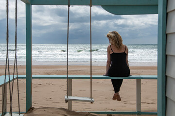 Blonde woman on the beach, sitting on the swing of a wooden house with the Atlantic Ocean in the...