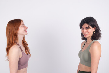 Two young women smiling and facing each other against a white background, expressing happiness and friendship.
