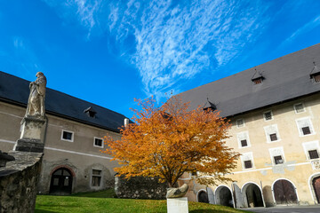 Exterior view of Benedictine monastery Saint Lambrecht Abbey in nature reserve Grebenzen, Styria, Austria. Religious landmark with rich unique architect features. Vibrant golden tree in courtyard - obrazy, fototapety, plakaty