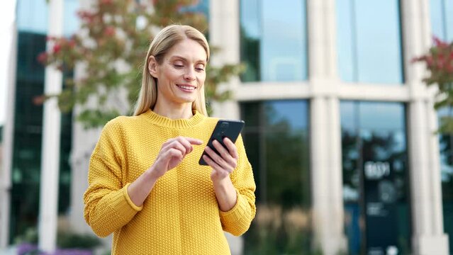 Happy woman is using the phone while walking on the street near a modern building. Smiling female tourist in a yellow dress reading a text message chatting online with friend or browsing social media