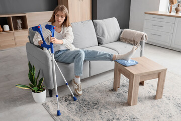 Injured young woman with crutches after accident sitting on sofa at home