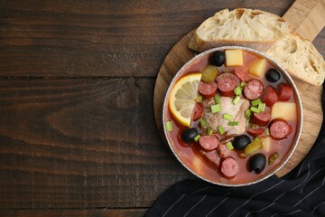 Meat solyanka soup with thin dry smoked sausages in bowl and bread slices on wooden table, flat lay