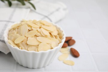 Fresh almond flakes in bowl on white tiled table, closeup. Space for text