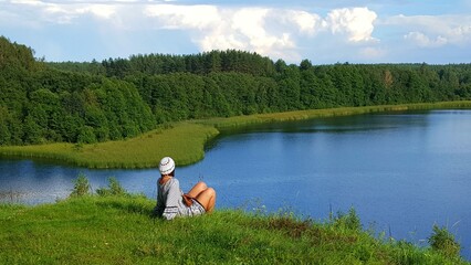 A girl admires the view of the lake from the top of a green hill. - obrazy, fototapety, plakaty