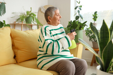 Senior gardener with plant and spray water bottle sitting on sofa at home