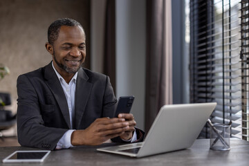 Confident African American businessman in a suit smiling while using his smartphone and laptop in a modern office setting.