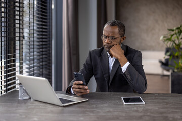 Serious businessman in suit working with smartphone and laptop at a modern office desk, focused and concentrated.