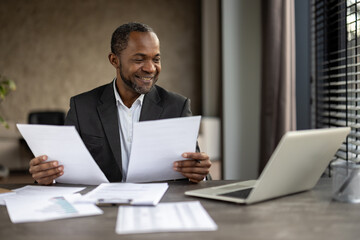 African American businessman in a suit smiles while reviewing papers in an office setting with a...
