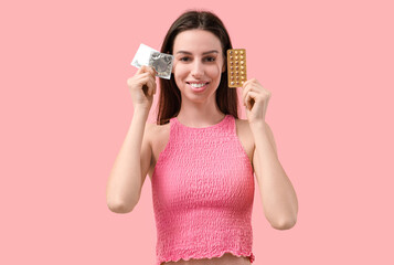 Beautiful young woman with birth control pills and condoms on pink background