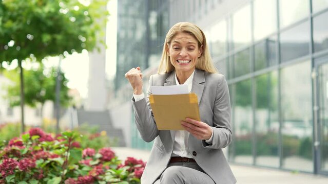 Excited businesswoman reading letter with great news sitting on a bench on the street near an office building. A satisfied woman is celebrating success, happy to receive a pleasant notification