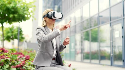 Businesswoman works using VR glasses in virtual reality simulator sitting on bench on street near office building. A woman uses gestures to control application programs and flips through virtual pages