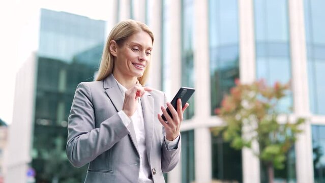 Smiling businessman is using a phone while walking on street near an office building. Happy mature female in a formal suit reading writes a message, chatting with a client or browsing online. Close up