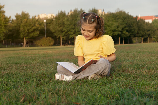 happy smiling cheerful girl 7 years old reads book, sits on grass in park and enjoys in summer day