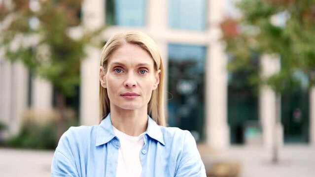 Portrait of an adult serious blonde woman with crossed arms standing on the street near a modern building. Head shot of confident mature female tourist in blue shirt posing looking at camera. Close up