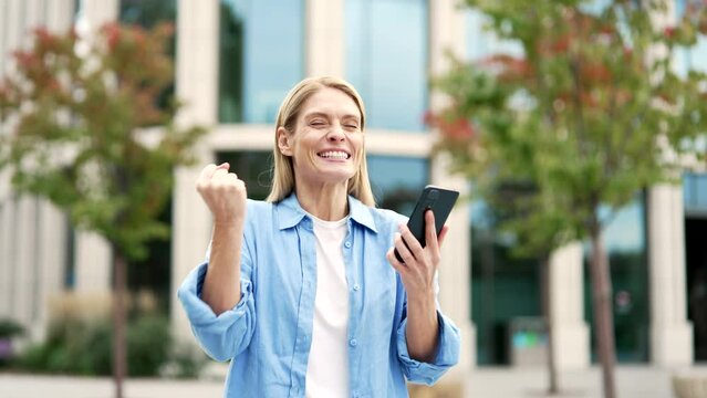 Excited woman is happy about great news she read on phone while standing on street near a modern building. Smiling joyful female celebrating success after receiving positive notification on smartphone