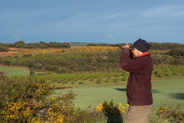 Man in profile looking through binoculars, dressed in mountain clothes, brown trousers, maroon...