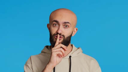 Middle eastern man doing hush silence symbol in studio, asking to keep a secret over blue...