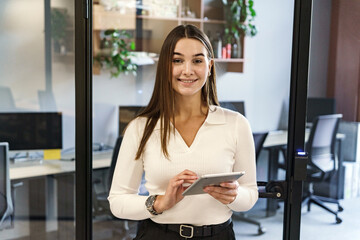 A poised businesswoman with a tablet stands at the threshold of an office, her welcoming smile and...