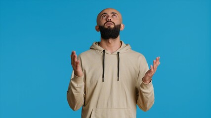 Naklejka premium Muslim person presenting his belief in Allah by praying on camera, looking up and saying a prayer for forgiveness. Middle eastern guy worshiping God with faith, religious man in studio. Camera 1.