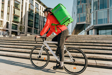 An urban courier in a vibrant red jacket and helmet expertly navigates his bicycle on city steps,...