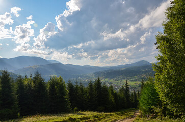 A well-trodden path leads into a valley of the Verkhovyna village nestled amidst rolling hills, adorned with lush greenery under the sky is painted with clouds filtering the golden sunlight