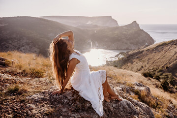 A woman in a white dress is sitting on a rock overlooking a body of water. She is enjoying the view and taking in the scenery.
