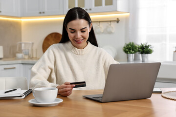 Happy young woman with credit card using laptop for shopping online at wooden table in kitchen