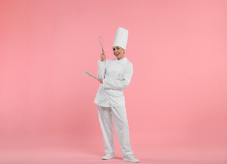 Happy professional confectioner in uniform holding whisk and spatula on pink background
