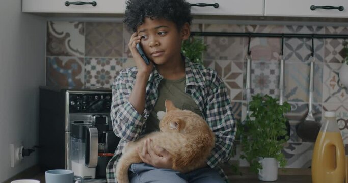 Cute African American boy speaking on mobile phone holding kitten sitting on kitchen table at home. Communication and childhood concept.
