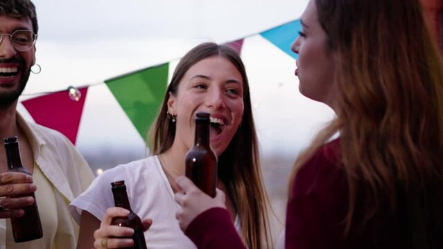 Focus on happy blonde woman with excited group of friends holding beer bottles at sunset rooftop party. Gathering of cheerful young people enjoying a summer day outdoors dancing and toasting lively