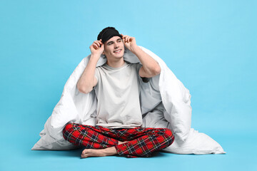 Happy man in pyjama and sleep mask wrapped in blanket on light blue background