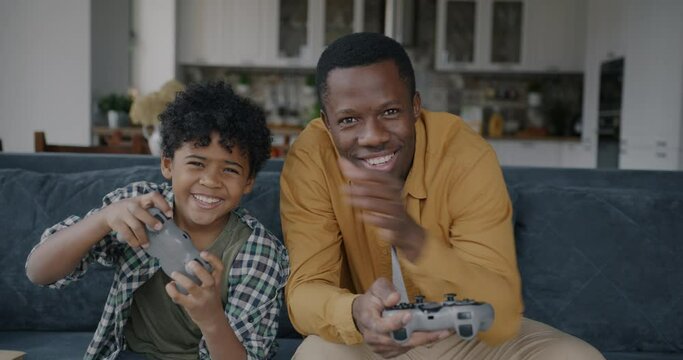 Slow motion portrait of excited family father and son playing video game with joysticks laughing having fun looking at camera. Modern lifestyle and happy childhood concept.