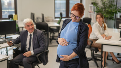 A happy pregnant woman stands in the middle of the office next to a Caucasian woman and an elderly...