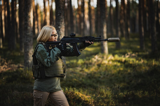 Blonde young military girl aims with a modern AK 12 rifle in the forest at war.