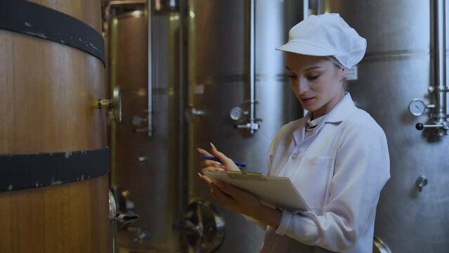 A focused female quality control specialist in a white coat inspects wine barrels in a fermentation cellar, clipboard in hand.
Keywords