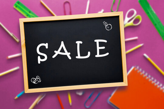 Stationery sale. Word sale is written with white chalk on a black school board on a blurred background of school supplies on a pink table. Template for advertising, online store.