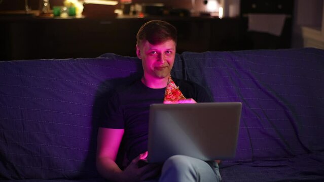 Portrait of cheerful young man watching content on laptop screen and eating pizza at night sitting alone on sofa. Happy male watching movie or online football game, relaxing after workday, slow motion