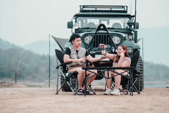 Man and woman share a cozy coffee break by a lake, with a rugged off-road vehicle and camping tent in the background.
