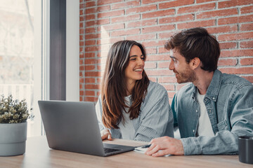 Happy young family couple bonding laughing using laptop looking at screen sit at table, smiling husband and wife buying choosing goods online doing internet shopping order with delivery at home.