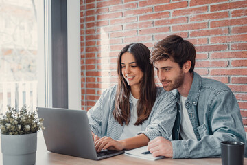Happy millennial couple sit at table at home browsing web on laptop shopping online together, smiling young husband and wife work on computer at desk pay household bills or taxes in internet banking.