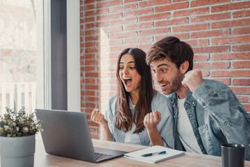 Overjoyed millennial couple sit at desk look at laptop screen feel euphoric with online win, excited happy young husband and wife read good news, get unexpected notification on computer, luck concept. - obrazy, fototapety, plakaty