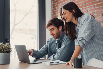 Happy young family couple bonding laughing using laptop looking at screen sit at table, smiling...